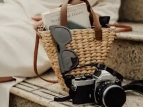A straw tote bag with a book peeking out, black sunglasses, and a vintage camera are placed on a stone step, suggesting travel essentials.